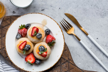 Cottage cheese pancakes, cheesecakes with fresh blueberries, strawberries, honey and mint leaves on a stylish wooden board on a gray concrete table. Natural products. Healthy and delicious breakfast.