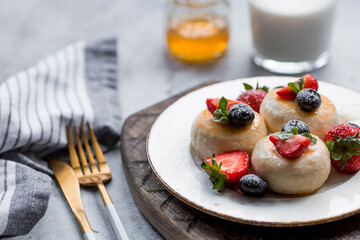 Cottage cheese pancakes, cheesecakes with fresh blueberries, strawberries, honey and mint leaves on a stylish wooden board on a gray concrete table. Natural products. Healthy and delicious breakfast.