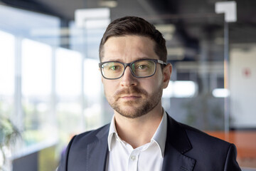 Fototapeta premium Portrait of young successful serious businessman, broker businessman close up in glasses and business suit looking at camera, workplace man working inside office, thinking about decision