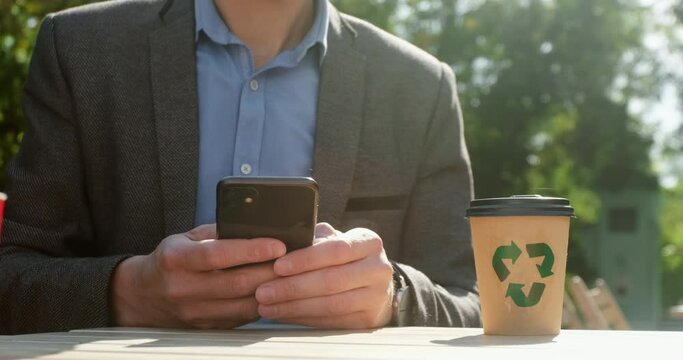 A busy man in a jacket quickly types on a smartphone, checks the time on his wrist watch, next to him is a glass of coffee with a recycling sign.