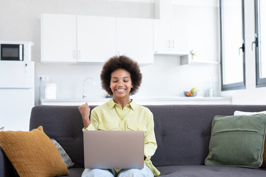 Excited Cheerful Young Black Woman Using Laptop Computer On Sofa At Home, Getting Good News, Feeling Joy, Dancing With Hands, Singing, Laughing, Making Winner Gesture, Happy To Win Prize.