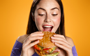 Close up of beautiful woman bites big delicious burger. Hungry girl eats hamburger with pleasure, looks with appetite at junk food, breaking diet, stands over orange background