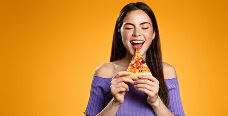 Excited 25 years woman bites tasty slice of pizza, orders delivery takeout from restaurant, hungry girl eats isolated on orange background