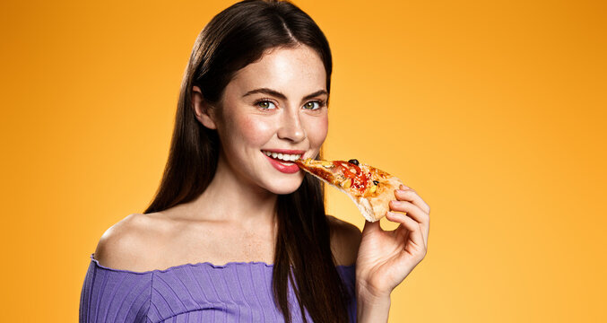 Photo of teenager enjoys delicious slice of pizza, likes this taste, closes eyes from pleasure, has good appetite, dressed in casual white shirt, isolated over yellow background. Hungry woman indoor