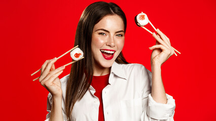 Enthusiastic woman with chopsticks, eats sushi rolls and laughs, orders delivery takeaway from asian restaurant. Girl enjoys asian food, japanese maki with salmon and tuna, red background