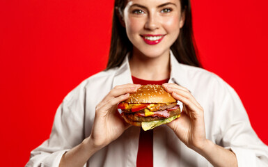 Portrait of smiling teen girl holds big tasty burger, eats hamburger and looks at camera, isolated on red background