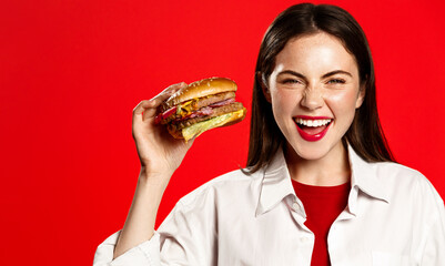 Enthusiastic young woman holds big tasty burger and laughs with joy, order takeaway, fast-food restaurant delivery, eats hamburger against red background