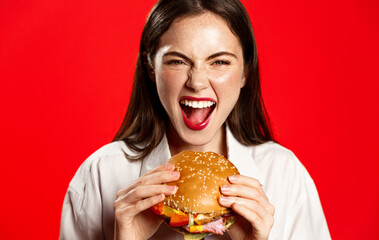 Cheerful pretty woman with a hamburger in the hands of a fast food diet, eats delicious burger on red background