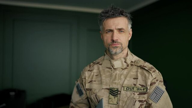 A soldier in camouflage stands with a laptop and looks at the camera
