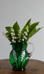Freshly cut, sweetly scented Lily of the Valley (Convallaria majalis) in an Antique Victorian green glass jug with white enamelling of the  flowers on an oak table. Portrait image with space for text. - 600440406