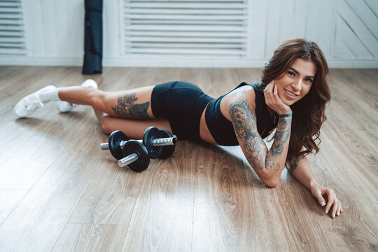 Dressed in black sportswera tattooed fitness girl poses lying on floor around dumbells in appartment.
