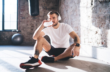 Confident sportsman in headphones drinking water while sitting on floor