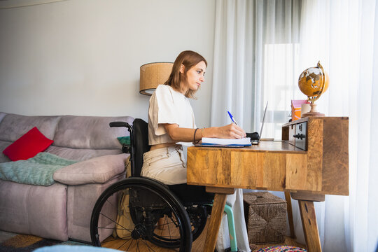 Young woman sitting on wheelchair while taking notes from laptop