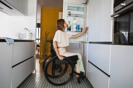Young disabled woman on wheelchair beside opened fridge in kitchen