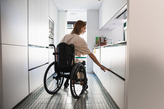 Young disabled woman on wheelchair beside opened cupboard in kitchen