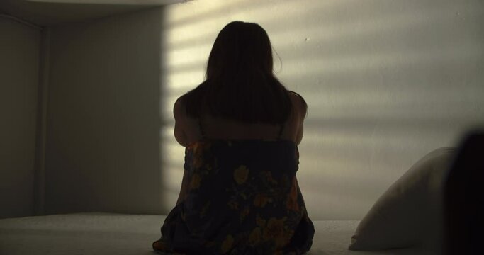 A lonely woman is sitting on the bed in a narrow dark room. This concept use for symptoms of stress, major depressive disorder (MDD), psychotic symptoms.