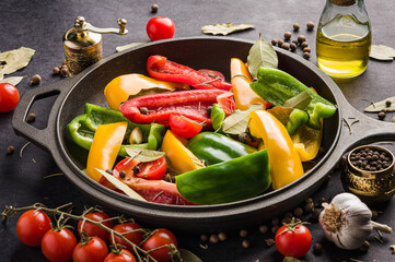 Vegetables, peppers and cherry tomatoes in a black frying pan with garlic, bay leaf and a small bottle of olive oil on a black background