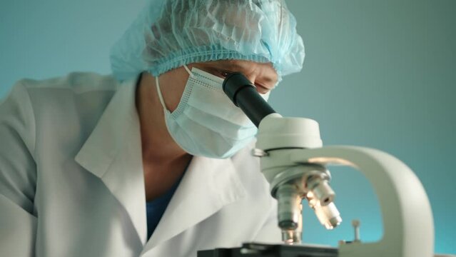 Medium shot of male middle-aged scientist conducting laboratory research on microscope in modern vet clinic. Doctor wears medical mask, hat, lab coat. High quality 4k footage