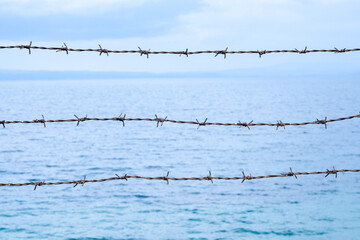 Close up barbed wire barrier against the sea and blue sky landscape. Metaphoric barbed wire is like...