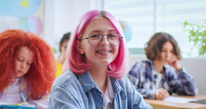 Happy teenage Caucasian girl in glasses and with pink hair sitting at desk and looking to the camera and smiling. Cheerful stylish female student answering at lesson. Educational concept.