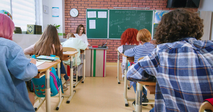 Back view of preteen students sitting on their personal desks at modern classroom. Confident pupils using laptops and tablets while doing exam or solving mathematical equations.