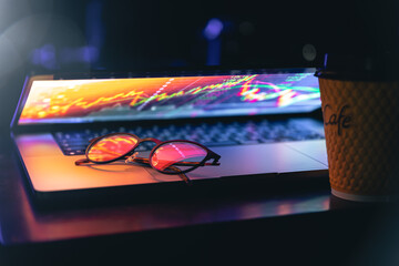 A laptop half closed in the dark with colourful glow, cup of coffee and glasses.