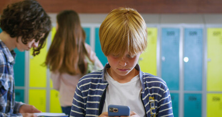 Portrait of caucasian schoolboy surfing internet on smartphone while standing on school corridor during break. Preteen boy relaxing after classes with modern gadget in hands.