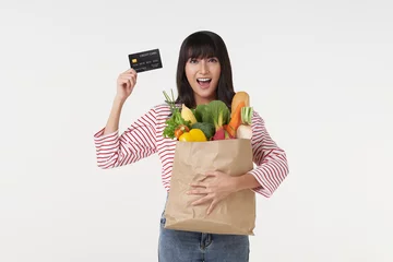 Zelfklevend Fotobehang smiling happy young asian housewife showing, presenting credit card and  holding paper bag with fresh food products isolated on wite background studio. shopping in supermarket © NaMong Productions