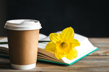 Paper cup, book and yellow flower on a blurred wooden background.