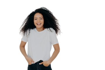 Half length shot of good looking smiling woman laughs at funny joke, has fun, keeps both hands in pockets of jeans, wears white t shirt, has curly fluffy hair, poses indoor, blank space on right side