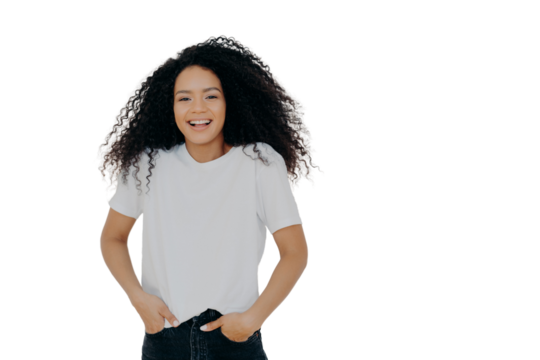 Half length shot of good looking smiling woman laughs at funny joke, has fun, keeps both hands in pockets of jeans, wears white t shirt, has curly fluffy hair, poses indoor, blank space on right side