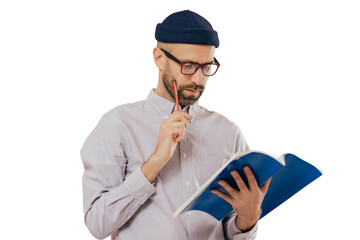 Concentrated unshaven adult man holds blue textbook and pencil, reads necessary information, has...