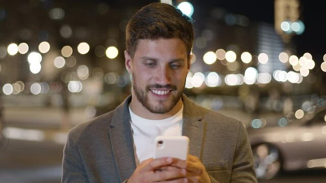 Lateral Dolly Close Up Slow Motion Successful And Cheerful Young Brunette Businessman Using Smartphone In City Financial District. He is Thinking, Sending Messages and Laughing. Traffic Night.
