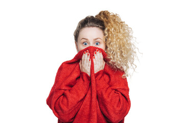 Frightened stunned female sees her phobia, covers face with collar of red sweater, stares at camera with scared look, isolated over white studio background. People, reaction and emotions concept