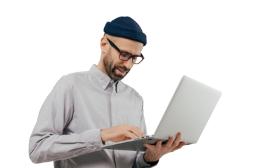 Concentrated bearded male administrative worker coordinates work on distance, searches information on laptop computer, wears white formal shirt and transparent glasses, poses indoor over grey wall