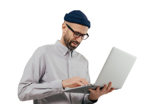 Concentrated bearded male administrative worker coordinates work on distance, searches information on laptop computer, wears white formal shirt and transparent glasses, poses indoor over grey wall