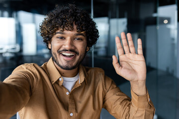 Smiling and cheerful businessman in office taking selfie photo on phone and talking on video call with colleagues and friends using smartphone, african american man waving at camera greeting gesture.