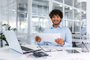 Portrait of young successful hispanic businessman inside office, man smiling and looking at camera, paper worker happy with achievement results sitting at workplace with laptop.