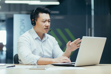 Fototapeta premium Young Asian man, sales consultant, support service agent in headset. Sits in the office at the laptop and consults, talks through a video call from the laptop.