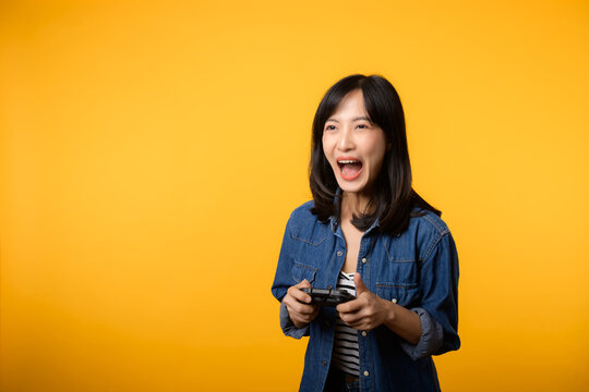 Portrait young asian woman with happy success smile wearing denim clothes holding joystick controller and playing video game. Fun and relax hobby entertainment lifestyle concept.