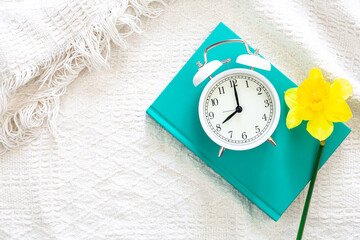 Book, alarm clock and flower in white bed, top view.