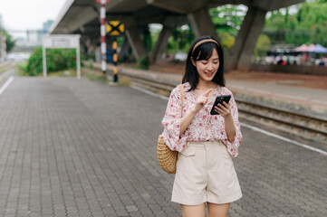 Fototapeta premium Asian young woman traveler with weaving basket using a mobile phone beside railway train station in Bangkok. Journey trip lifestyle, world travel explorer or Asia summer tourism concept.