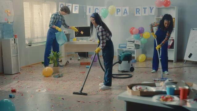 A multinational group of cleaners clean up the office after a corporate party. An African American woman, an Asian woman and a European man mop floors, vacuum and collect garbage in a trash bag.