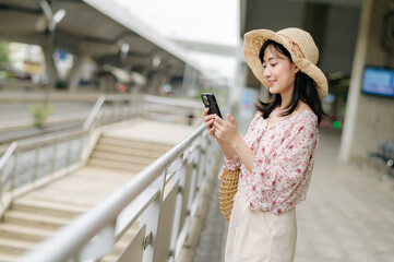 Asian young woman traveler with weaving basket using a mobile phone beside railway train station in Bangkok. Journey trip lifestyle, world travel explorer or Asia summer tourism concept.