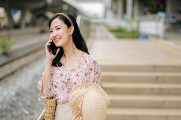 Fototapeta premium Asian young woman traveler with weaving hat using a mobile phone beside railway train station in Bangkok. Journey trip lifestyle, world travel explorer or Asia summer tourism concept.