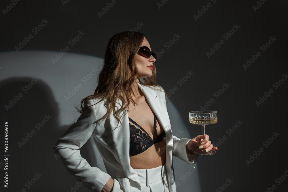 Wall mural Cool fashion beauty sexy woman with stylish sunglasses in a fashion white suit with a blazer and black lace bra with a vintage glass of champagne on a dark background in the studio. Pretty chic lady