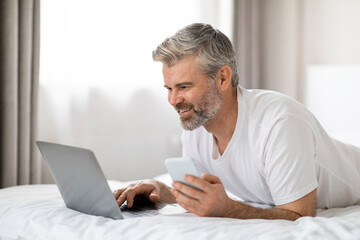 Happy grey-haired man lying on bed with phone and laptop
