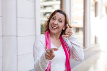 middle age woman smiling cheerfully and pointing to camera while making a call you later gesture, talking on phone