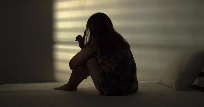 A lonely woman is sitting on the bed in a narrow dark room. This concept use for symptoms of stress, major depressive disorder (MDD), psychotic symptoms.