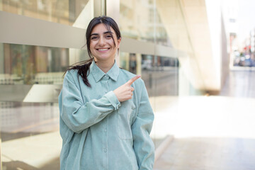 pretty woman smiling cheerfully, feeling happy and pointing to the side and upwards, showing object in copy space